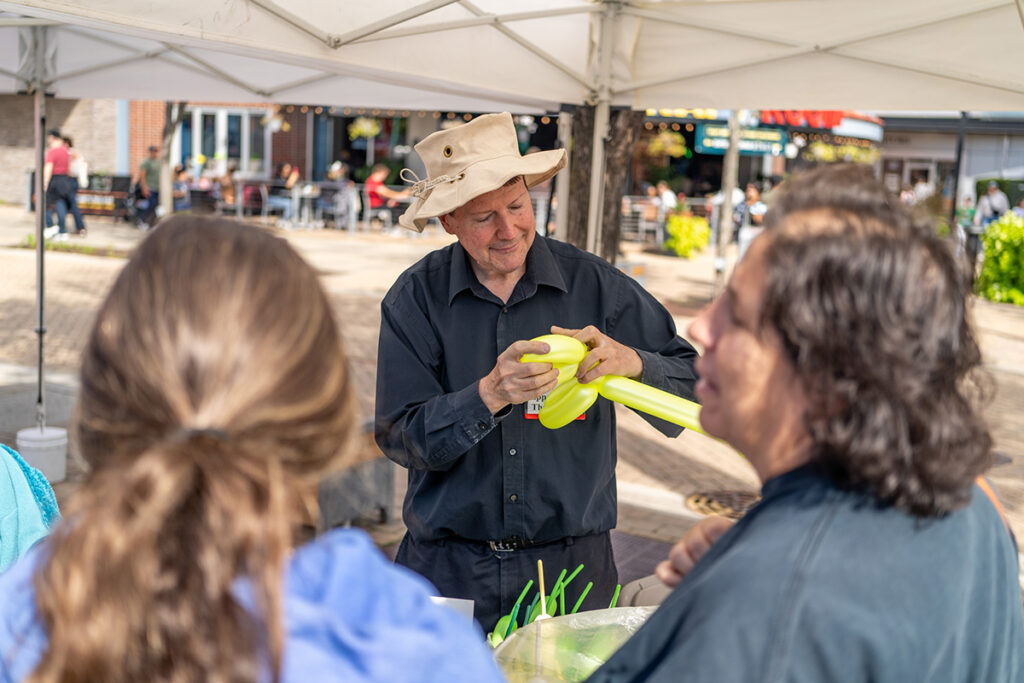 A man in a beige hat shapes a yellow balloon under a white canopy, while two people watch in the foreground at an outdoor event. Tables and people are visible in the background.