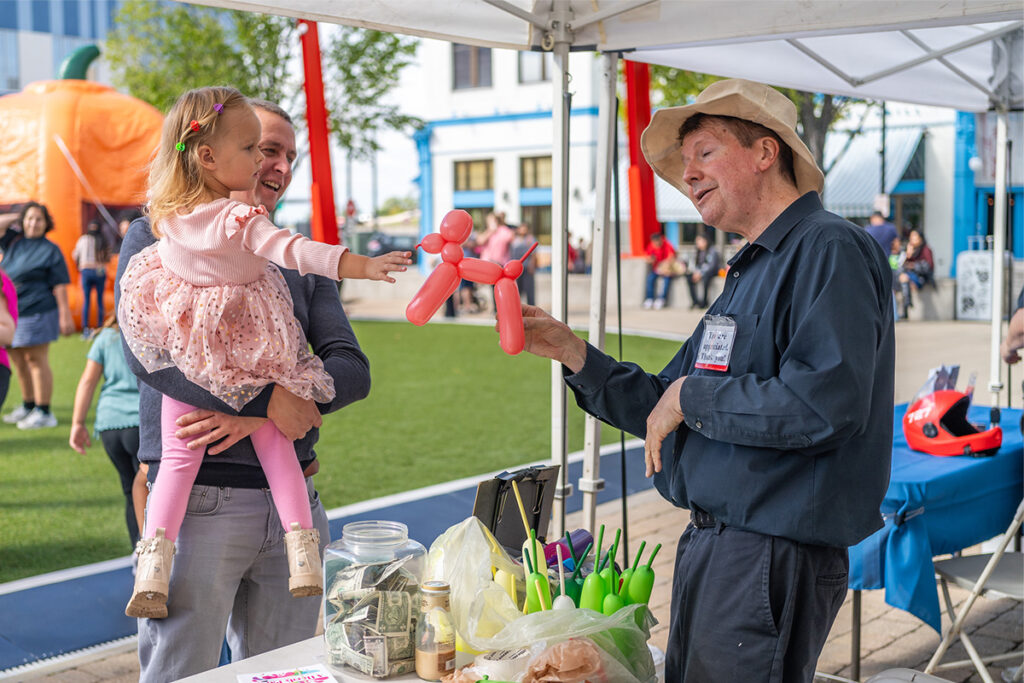 A man in a hat hands a pink balloon animal to a smiling little girl held by an adult at an outdoor fair. A table with cash, balloons, and supplies is in the foreground; people and booths are visible in the background.