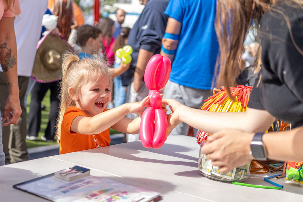 A smiling young girl reaches excitedly for a pink balloon animal being handed to her by an adult at a busy outdoor event with people and colorful balloons in the background.