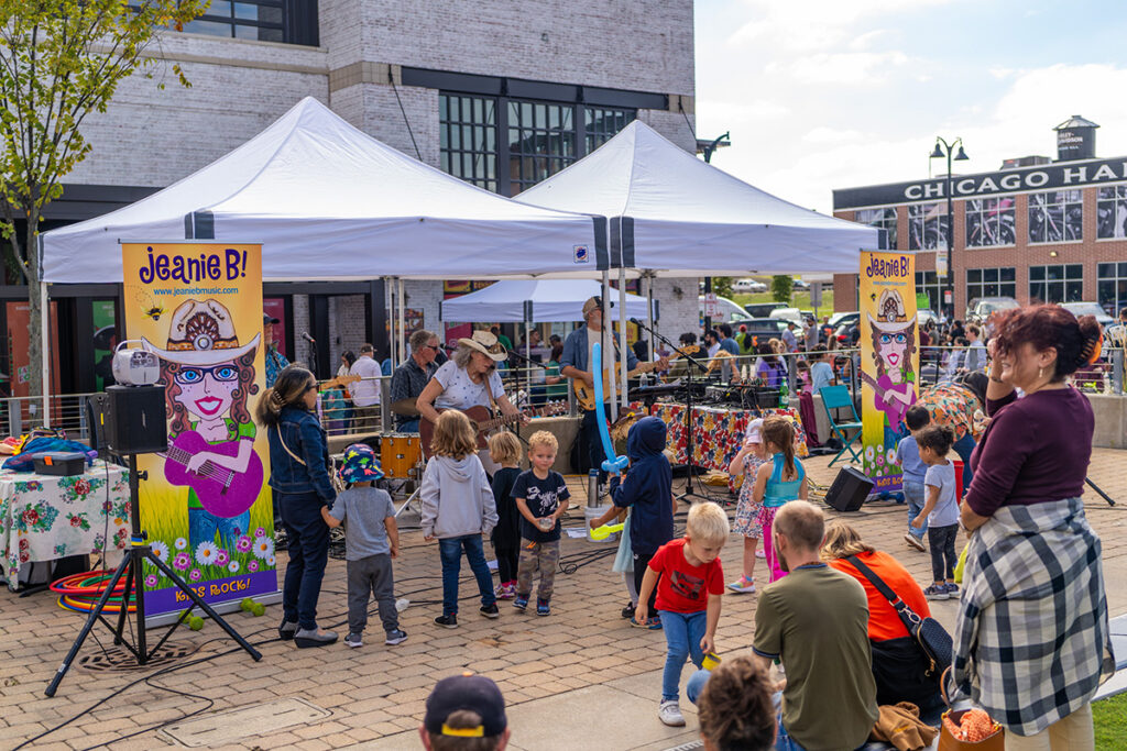 Children and adults gather outdoors near white tents for a live music performance by Jeanie B! Colorful posters and musical instruments are visible, with kids dancing and playing in front of the stage.