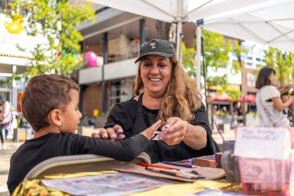 A smiling woman in a black apron and cap paints a young boy’s hand at an outdoor booth. Art supplies are on the table, and people walk by in the background under tents and trees.