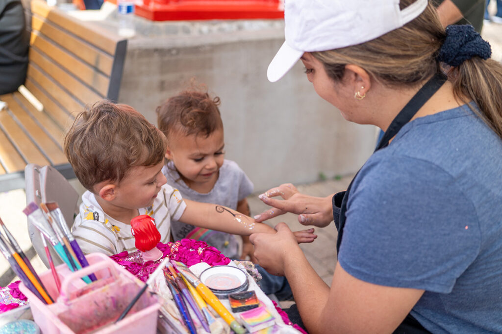 A woman wearing a white cap is face painting a young boy’s arm while another child looks on. Art supplies and brushes are on the table in front of them, and they are outdoors near a bench.