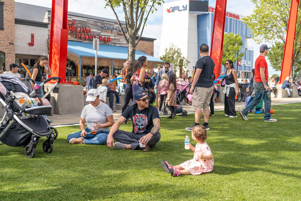 People enjoy a sunny day at an outdoor event. Some sit on the grass, including a man in sunglasses and a toddler. Others walk by with strollers. There are red banners, trees, and shops in the background.
