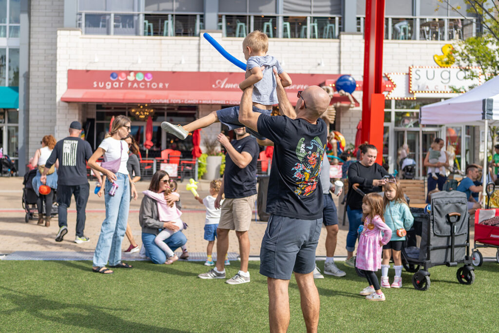 A man lifts a young child holding a balloon sword in an outdoor plaza, surrounded by families and children. People are gathered near shops and restaurants on a sunny day.