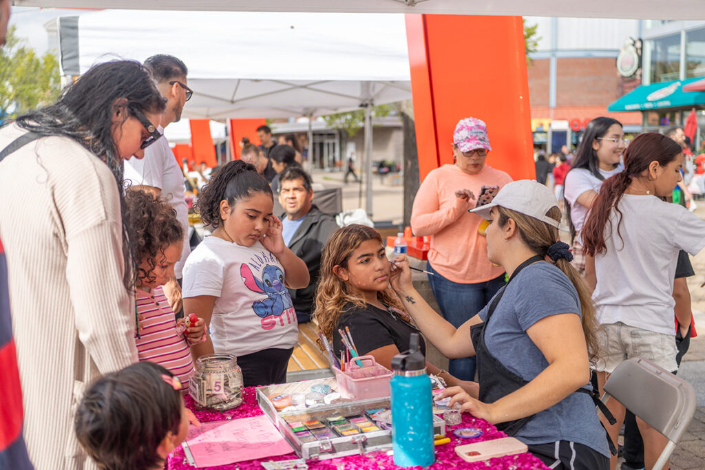 A group of people, including children, gather at a face painting booth outdoors. A woman paints a girls face while others wait and watch. The scene is lively with colorful decorations and a sunny atmosphere.