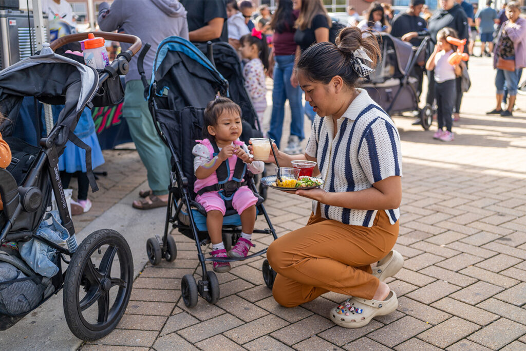 A woman kneels beside a stroller, offering food to a young child at an outdoor event. People and other strollers are visible in the background. The woman holds a plate and cup while the child reaches for food.