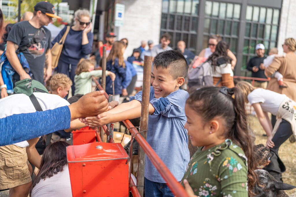 Children smile and reach out to pet animals at an outdoor petting zoo. Other kids and adults stand nearby, enjoying the sunny day. The scene is lively and cheerful, with a crowd gathered in the background.