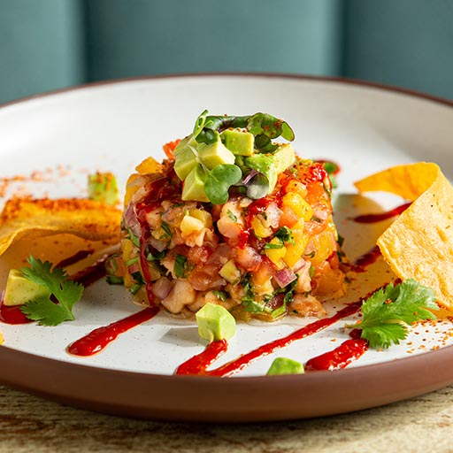 A colorful serving of ceviche topped with diced avocado, surrounded by tortilla chips, cilantro, and drizzled with vibrant red sauce on a white plate.