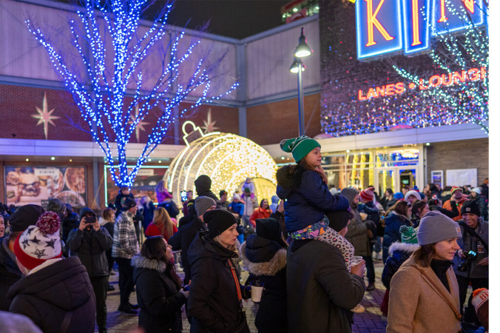A festive crowd gathers outdoors at night in winter clothing, with holiday lights, a large ornament decoration, and a child on an adult’s shoulders near a bowling alley entrance decorated with sparkling lights.