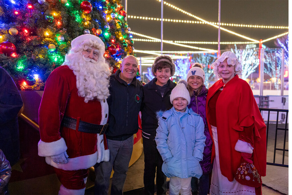 A family poses with Santa Claus and Mrs. Claus in front of a large, brightly lit Christmas tree at an outdoor holiday event at night. Everyone is dressed warmly and smiling.
