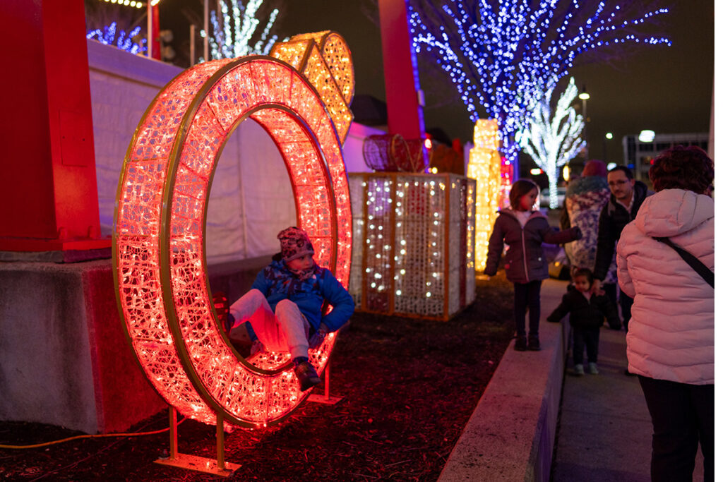 A child sits inside a large, illuminated red ring decoration at an outdoor light display, while adults and other children stand nearby. Trees are decorated with blue and white lights in the background.