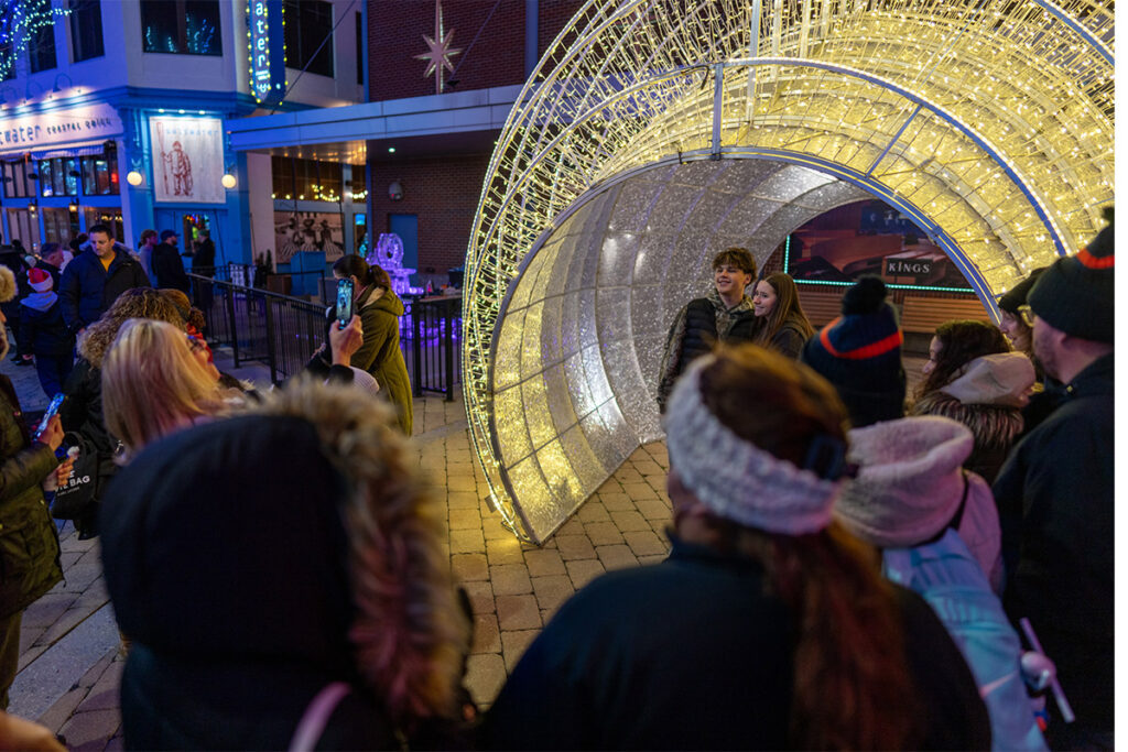 A crowd gathers at night around a glowing tunnel of holiday lights, while a woman takes a photo of two people posing inside the illuminated structure. Nearby buildings are decorated with festive lights.