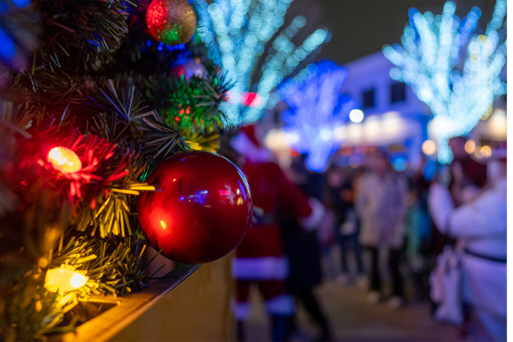 A close-up of a decorated Christmas tree with colorful lights and a red ornament, with blurred people and bright, blue-lit trees in the background at a nighttime outdoor event.