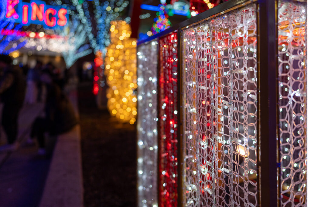 A festive outdoor scene with colorful holiday lights, including strands of white and red lights on a fence and blurred yellow and blue lights in the background. People are visible sitting and walking.
