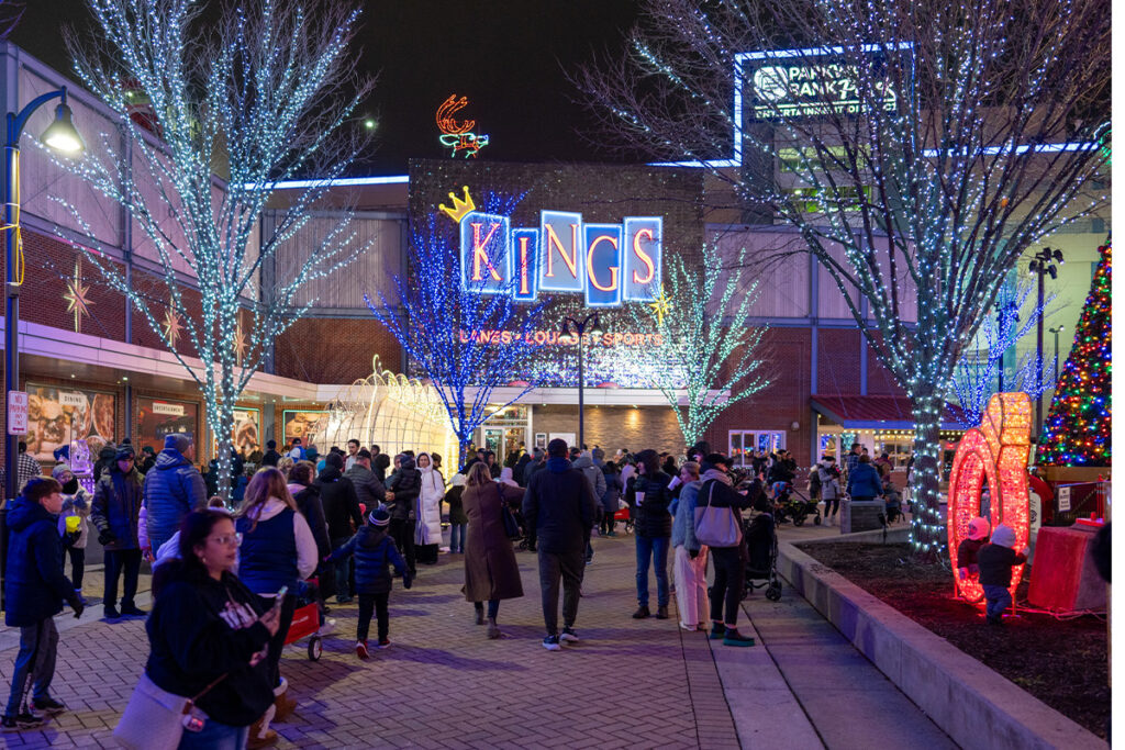 A crowd gathers at night in a festive outdoor plaza decorated with colorful holiday lights. A large “KINGS” sign and illuminated trees are visible, along with holiday displays and people enjoying the atmosphere.