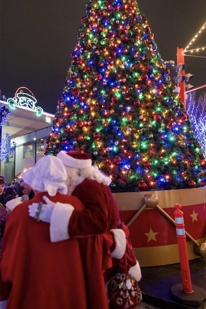 Mr. and Mrs. Claus hug in front of a large, brightly lit Christmas tree decorated with colorful lights during a festive outdoor event at night.