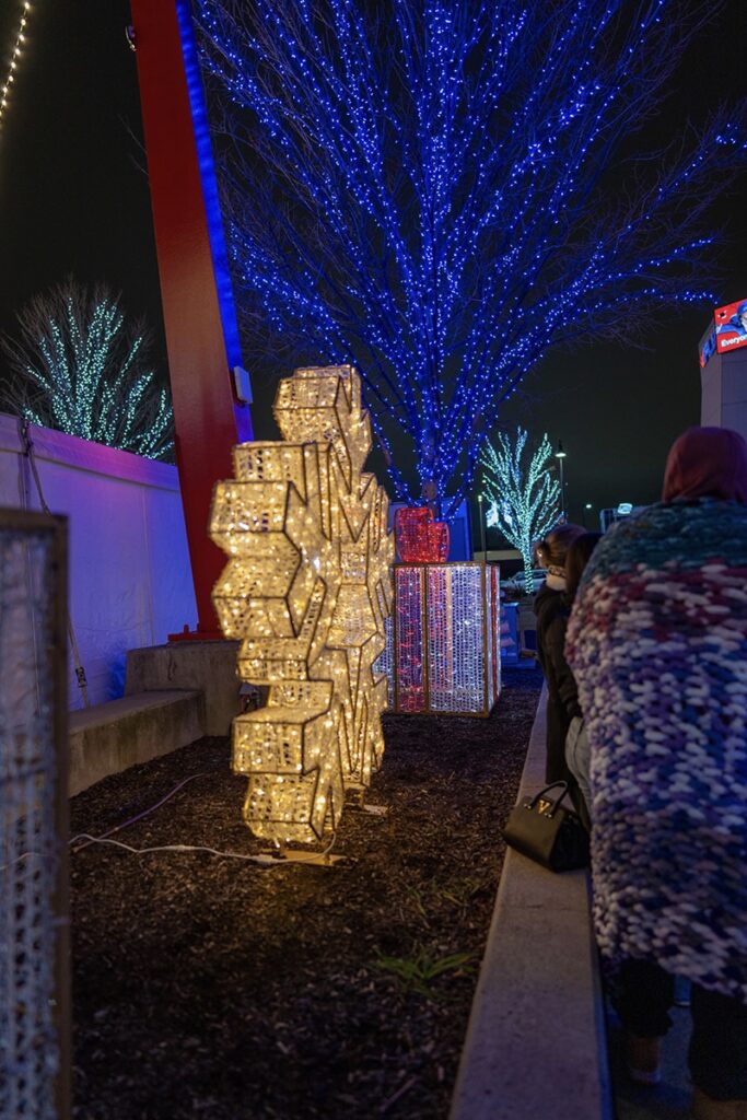 Large illuminated snowflake and rectangular decorations glow at night beside a walkway, with people in warm clothing nearby and trees wrapped in blue and white string lights in the background.