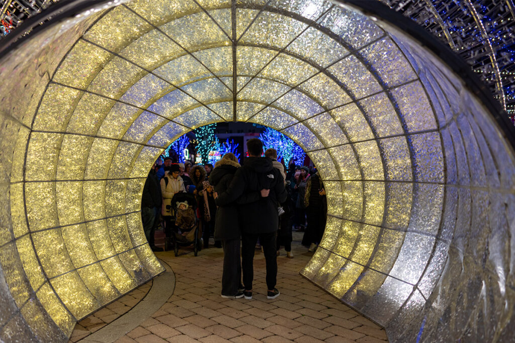 A couple stands arm in arm inside a glowing tunnel of holiday lights, looking out at a festive crowd and colorful illuminated decorations in the background.