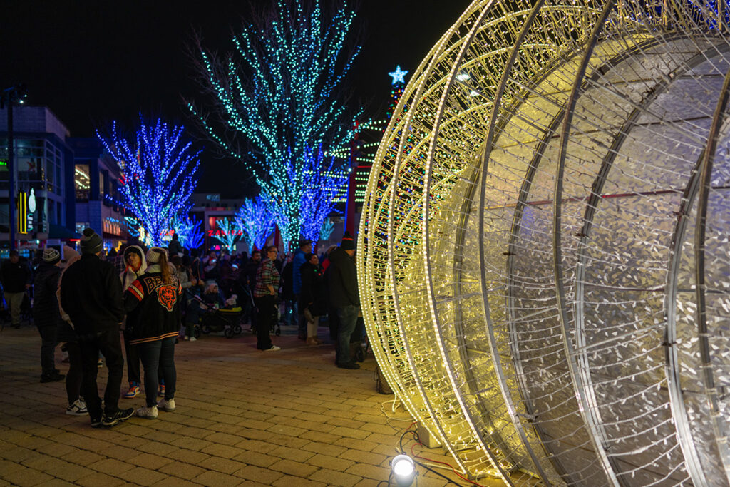 People gather outdoors at night near a large, lit holiday decoration and blue-lit trees. The area is festive, with lights illuminating the scene and people dressed warmly in jackets and hats.