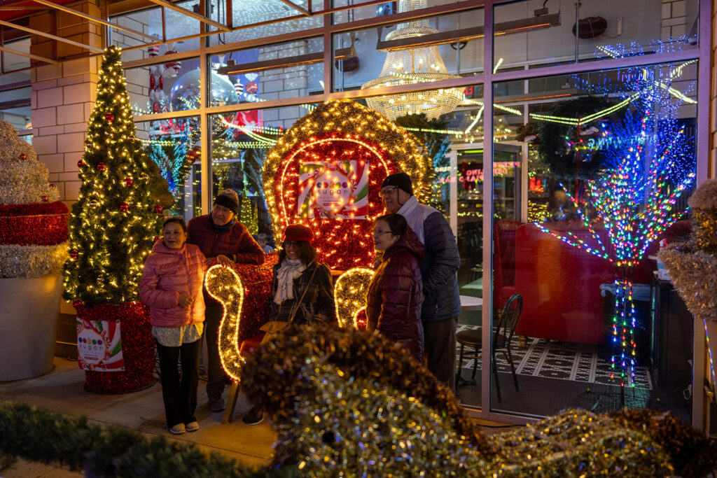 Five people pose and smile in front of a brightly lit holiday display with Santa’s sleigh and festive decorations, surrounded by colorful Christmas lights and ornaments outside a store or café at night.