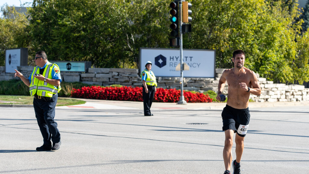 A shirtless male runner in black shorts passes through an intersection near a Hyatt Regency sign, while two police officers direct traffic and monitor the area on a sunny day.