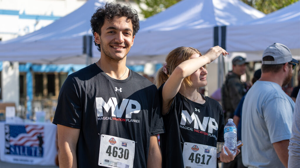 Two young runners wearing black MVP shirts and race bibs stand outside at an event. The man smiles at the camera while the woman beside him shields her eyes from the sun and holds a water bottle. White tents are in the background.
