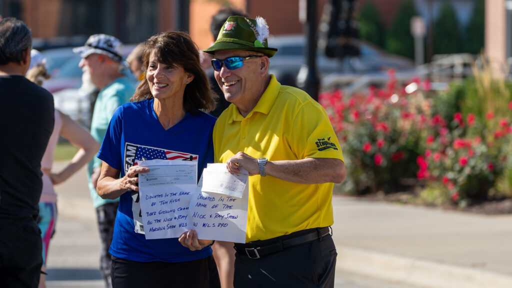 A smiling woman in a blue shirt and a man in a yellow shirt and green hat hold papers together while posing outdoors, surrounded by other people and blooming red flowers.
