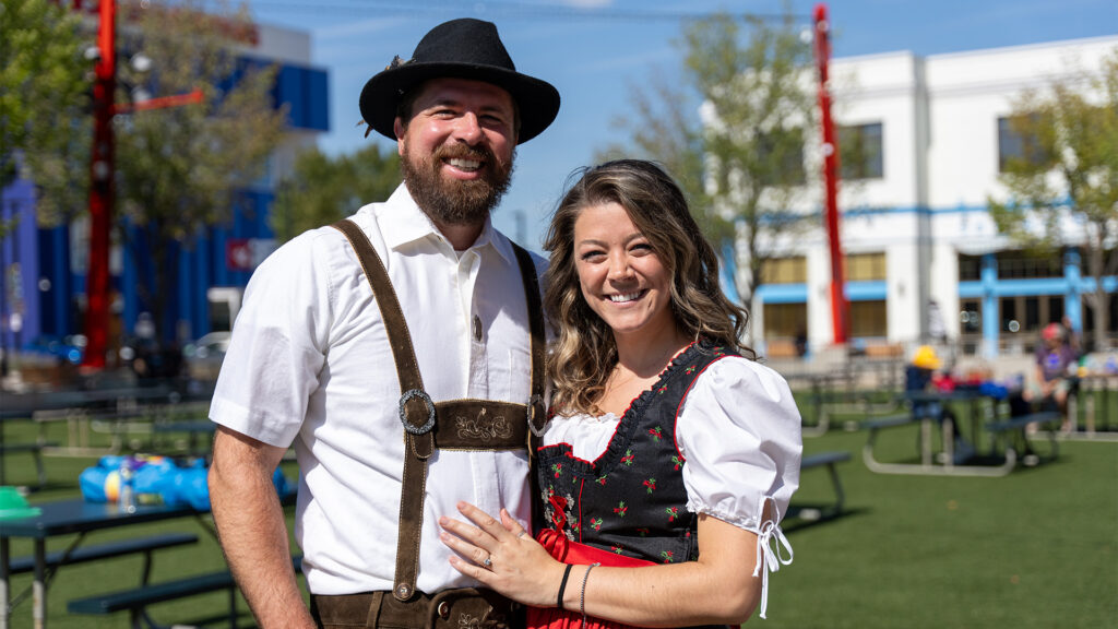 A smiling man and woman wearing traditional Bavarian clothing stand outdoors on a sunny day, with picnic tables and buildings in the background.