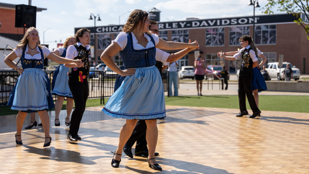 A group of people in traditional Bavarian costumes dance outdoors on a wooden floor, with a Harley-Davidson building and bystanders visible in the background on a sunny day.