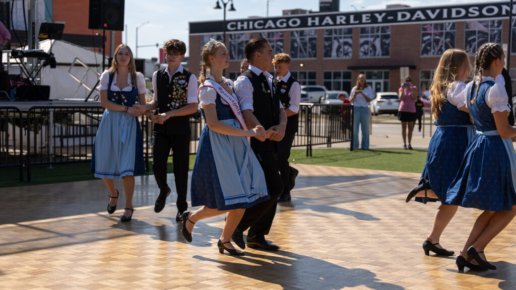 A group of young people in traditional German attire dance together on an outdoor wooden floor, with a Chicago Harley-Davidson building visible in the background.