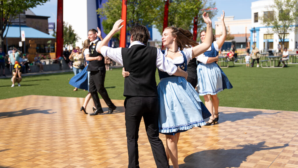 People in traditional Bavarian clothing are dancing on an outdoor wooden floor, with others and green grass visible in the background on a sunny day.