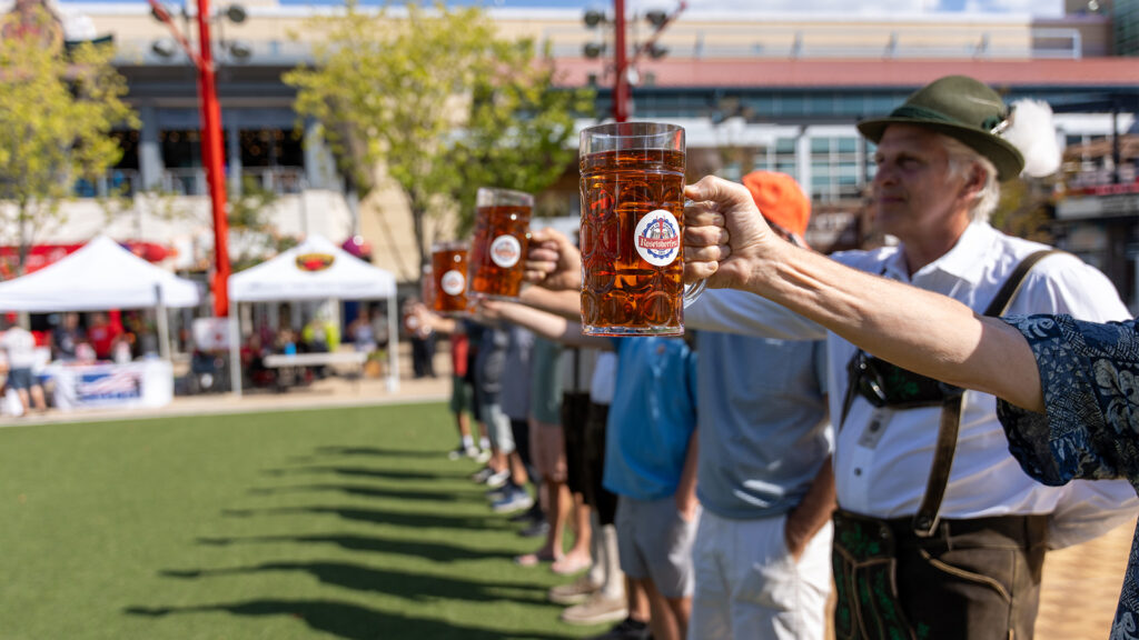A row of people, some in traditional German attire, hold large mugs of beer at arm’s length outdoors during a sunny festival, with tents and trees in the background.