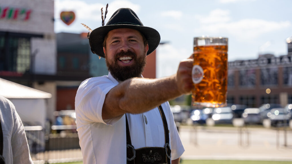 A smiling man wearing traditional Bavarian clothing and a hat holds out a large glass of beer outdoors on a sunny day, with buildings and cars in the background.
