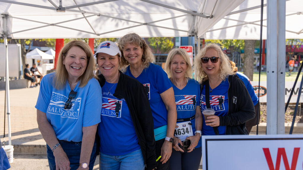 Five smiling women wearing blue SALUTE, INC. T-shirts and sunglasses stand together under a white tent at an outdoor event on a sunny day.