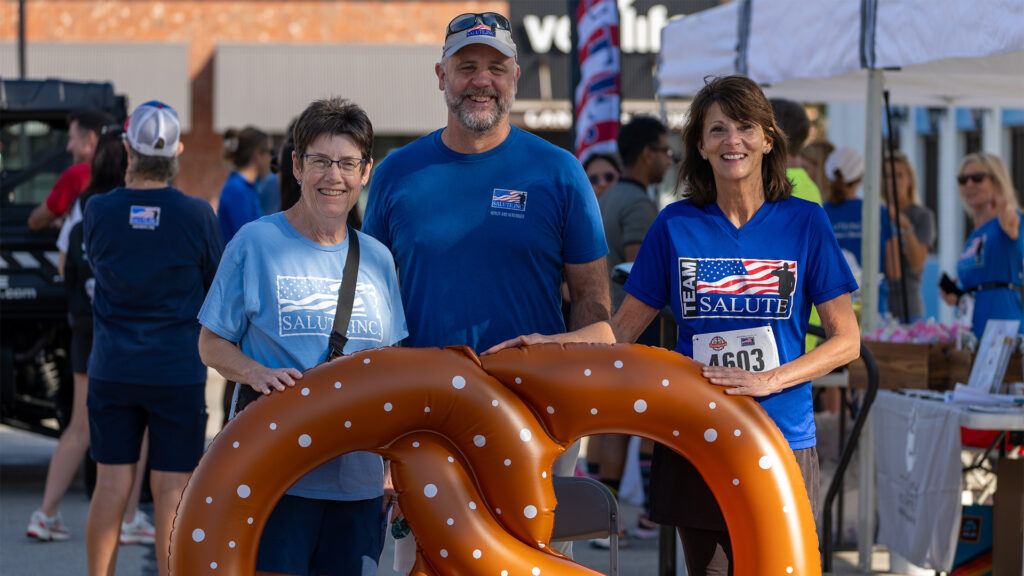 Three people wearing blue SALUTE shirts smile and pose with a large inflatable pretzel at an outdoor event, surrounded by other participants and tents in the background.