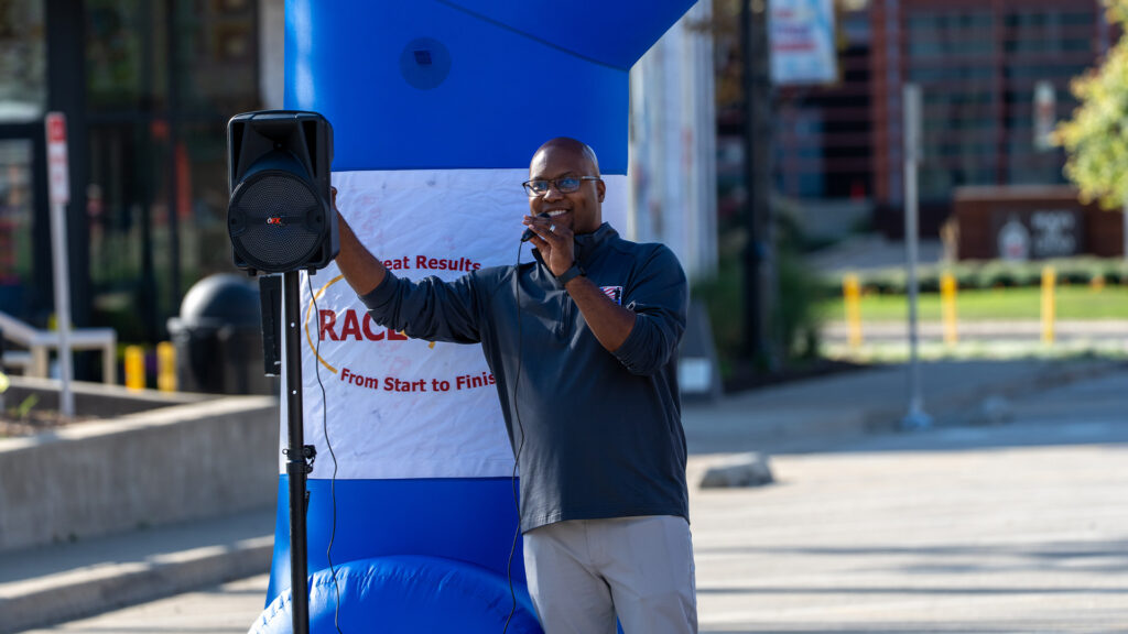 A man stands outdoors speaking into a microphone next to a blue inflatable arch and a speaker, with a RACE banner behind him. The background shows a sidewalk, buildings, and trees.