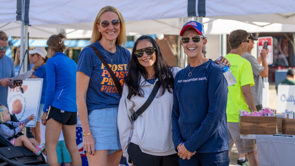 Three women smiling and posing together at an outdoor event with tents and other people in the background. One woman holds a water bottle, and they all wear casual clothes and sunglasses.