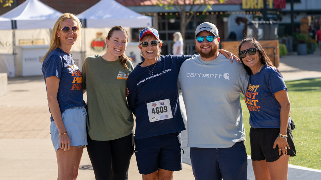 Five people stand outdoors smiling for a group photo. The person in the center wears a race bib and hat. The group is casually dressed, and a sunny event with tents and signs is visible in the background.