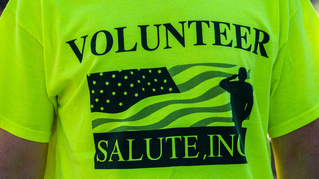A person wearing a bright yellow T-shirt with the text VOLUNTEER above an American flag graphic and a silhouette of a saluting figure, and SALUTE, INC written below.