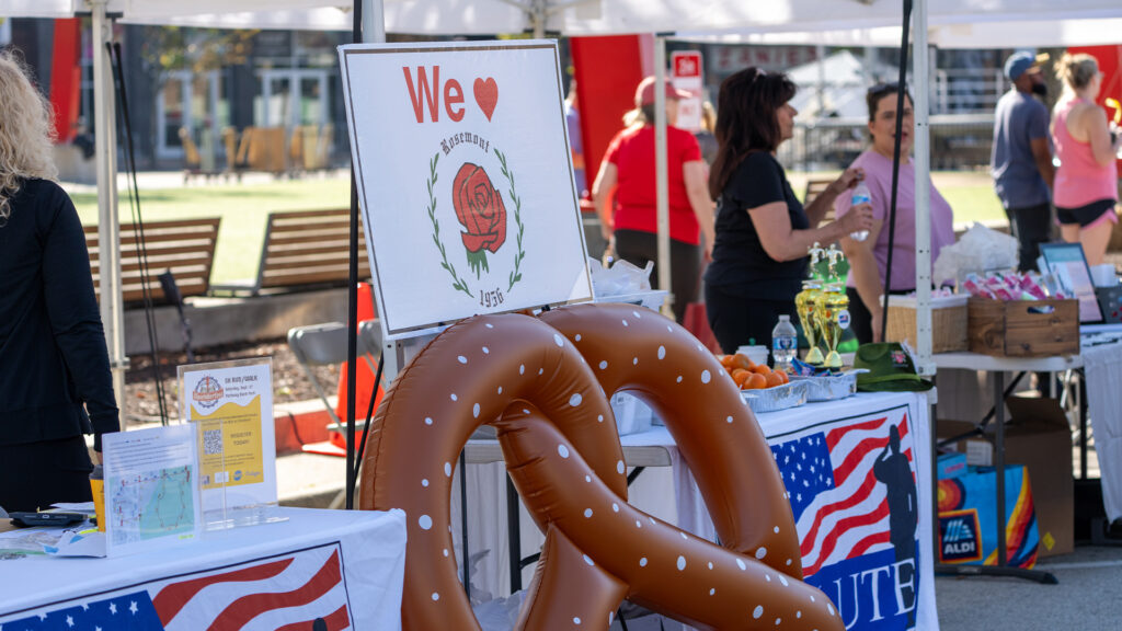 A booth at an outdoor event features a large inflatable pretzel and a sign with a rose and the words “We ♥” beside it. People stand behind the booth, which is decorated with American flag-themed tablecloths.