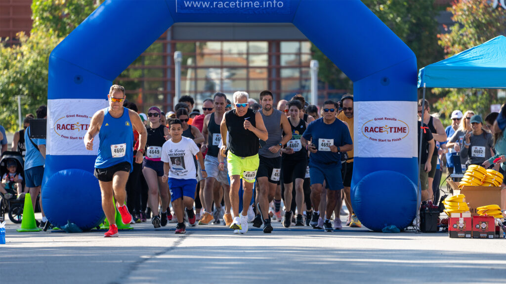 A diverse group of runners of all ages begin a race under a blue inflatable arch on a sunny day, with spectators and volunteers watching in the background.