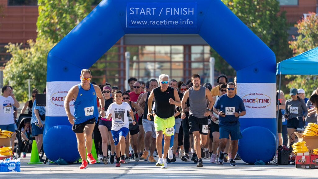 A group of runners, including adults and children, starts a race under a blue arch labeled START / FINISH on a sunny day, with trees and buildings visible in the background.