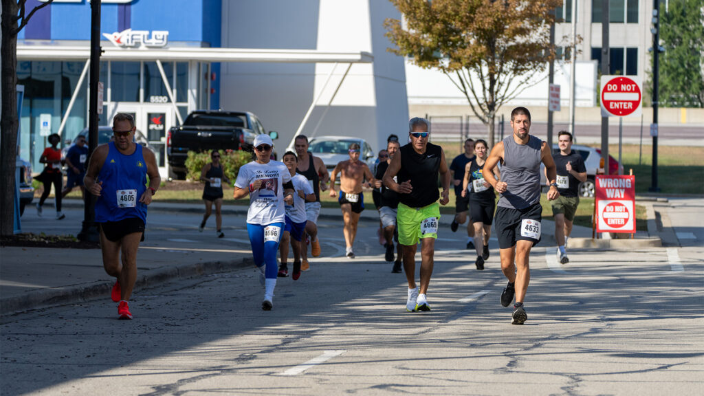 A group of runners, wearing race bibs, participate in a road race on a sunny day. Trees, signs, and a building with iFLY signage are visible in the background.