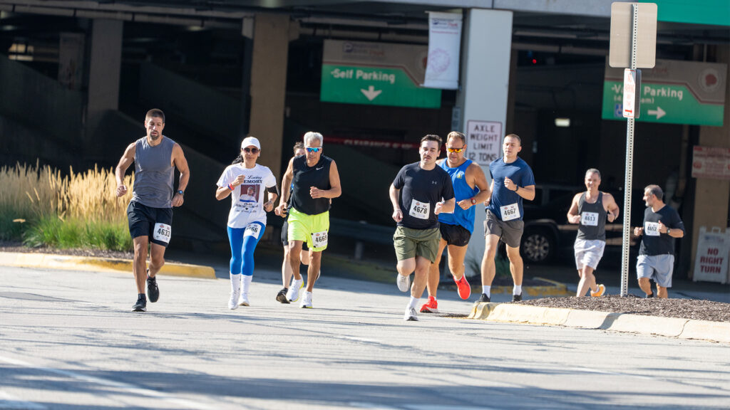 A group of runners wearing numbered bibs participate in a race on a sunlit street near a parking garage. Some runners wear sunglasses and hats, and all appear focused and determined.
