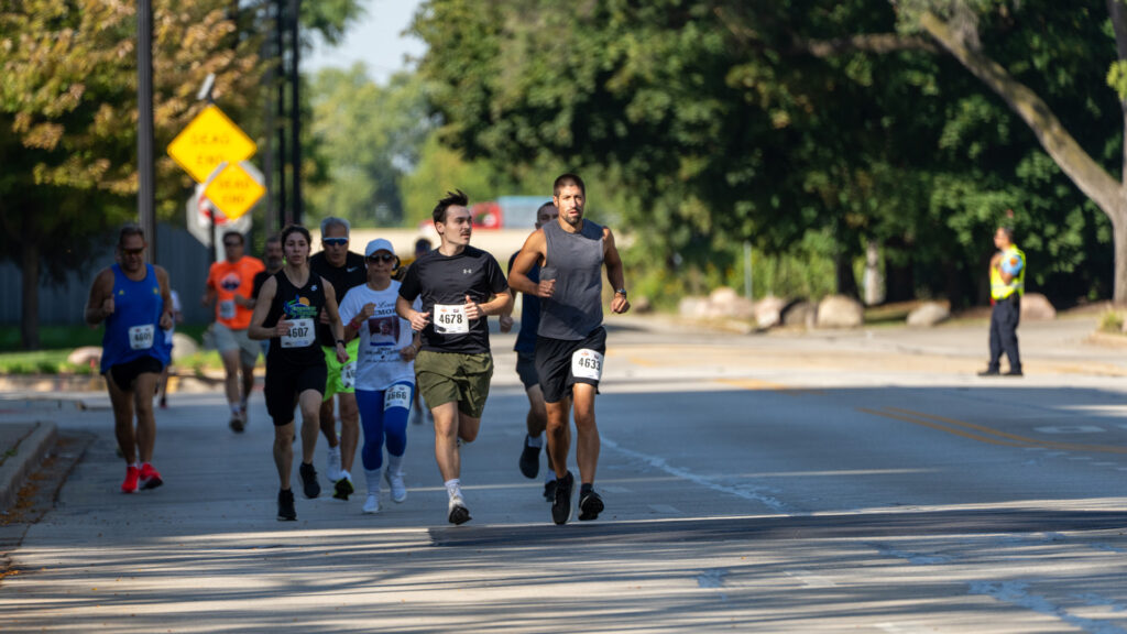 A group of runners wearing race bibs participate in a road race on a sunny day, with trees and a police officer visible in the background. Two leading runners are in the foreground.