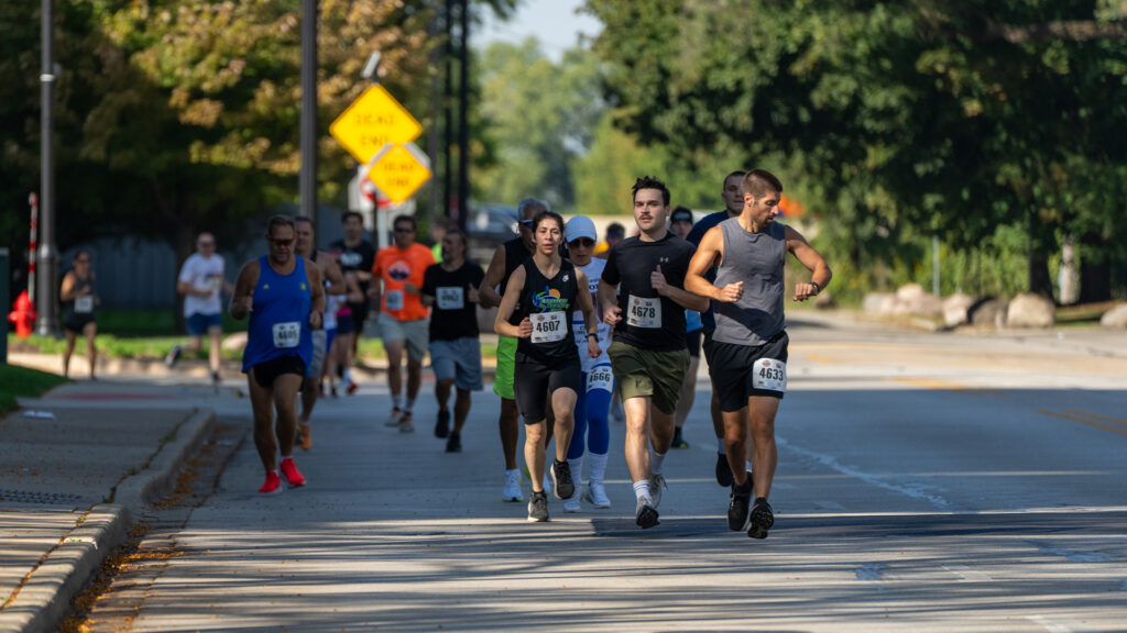 A group of runners with race bibs jog on a sunny street during a road race, with trees and road signs visible in the background. The leading runners appear focused and determined.