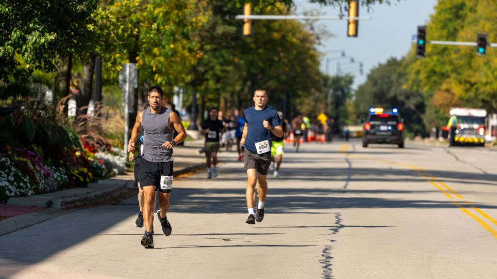 Two men run on a sunny street during a race, wearing numbered bibs. Other runners are visible behind them. Police and emergency vehicles are parked ahead, and trees line both sides of the road.