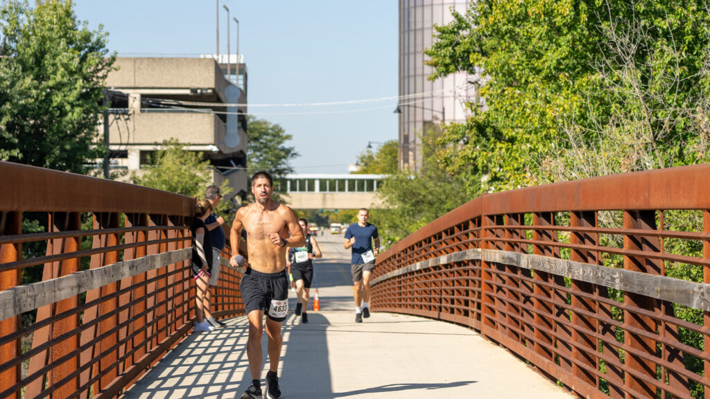 A shirtless man runs across a pedestrian bridge during a race, with several other runners following behind him. Trees and buildings line the sides under a clear, sunny sky.