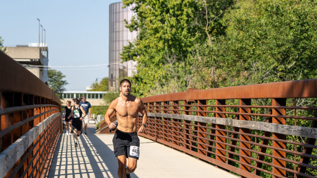A shirtless man wearing black shorts and a race bib runs across a bridge, with other runners following behind. The bridge is rusty orange and surrounded by green trees and buildings in the background.