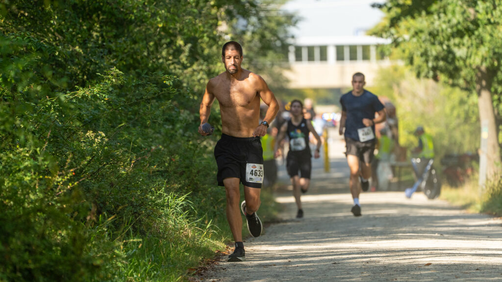 A shirtless man with a race bib jogs on a sunlit dirt path surrounded by trees, with several other runners behind him, participating in a race.